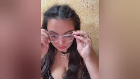 A video of a woman with long dark hair, light brown skin, and large breasts, wearing a black lace bra and clear glasses, adjusting them against a textured beige wall.