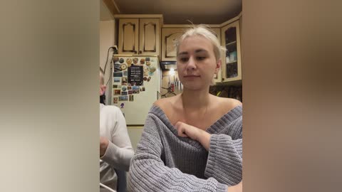 Video of a young woman with blonde hair, wearing a grey off-shoulder sweater, sitting in a cozy kitchen with wooden cabinets and a refrigerator covered in magnets.