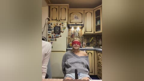Video of a woman in a gray sweater, red blindfold, seated in a wooden kitchen, with beige cabinets and a colorful fridge.