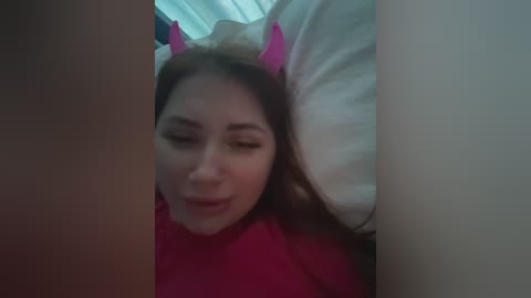 A video of a young woman with light skin, wearing a pink devil costume with horns, lying on a bed with white pillows.