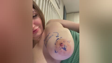 Video of a topless woman with fair skin, medium-sized breasts, and a tattoo on her right breast. She has long brown hair and is smiling. Background shows a white room with vertical blinds and a green blanket.