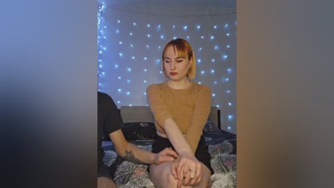 Video of a young woman with short, red hair, wearing a mustard-yellow top, sitting on a bed with a man's hand on her leg in a dimly lit room with string lights.