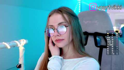 Video of a young Caucasian woman with long, straight blonde hair and fair skin, wearing round glasses and a white top, sitting in a gray gaming chair in a brightly lit room with blue LED lights.