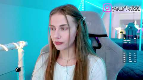 Video of a young, fair-skinned woman with long blonde hair, wearing a white shirt, seated in a gaming chair. Background features a glowing blue light, gaming accessories, and social media watermark.