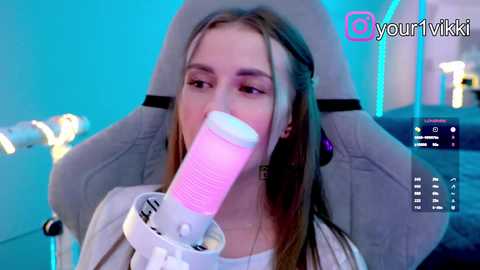 Video of a young woman with long brown hair, wearing a headset and white top, sitting in a gray gaming chair. She is inhaling from a pink disposable vape pen. Background includes a glowing blue light and a computer screen displaying a game.