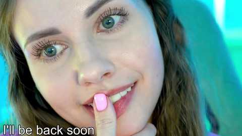 Video of a young woman with fair skin, green eyes, and long brown hair, smiling and biting her pink nail. Background is a blurred blue-green gradient. Text reads \"I'll be back soon.\