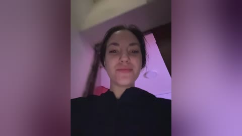 A video of a young woman with fair skin, dark hair in braids, wearing a black shirt, smiling slightly. Background features a pinkish-purple tint, and a white ceiling with a circular light fixture.