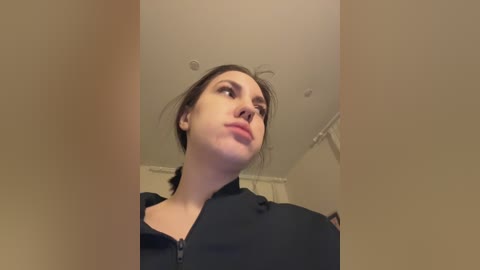 A video of a young woman with fair skin and dark hair, wearing a black jacket, standing indoors against a beige wall. She looks upward, with a neutral expression.