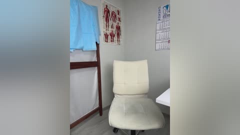 Video of a minimalist medical examination room featuring a beige office chair, a wooden examination table with a blue towel, anatomical posters on gray walls, and a white desk partially visible.