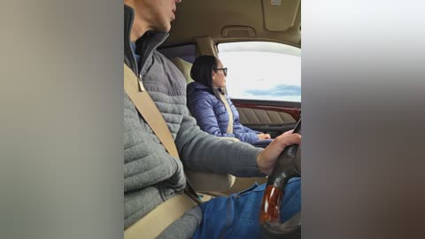 Video of a woman in a grey puffer jacket and a man in a blue jacket driving a car. Both are in the front seats, looking ahead.