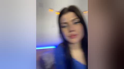 A blurred video of a young woman with fair skin, dark hair, and wearing a blue top, possibly in a nightclub setting with blue lighting.