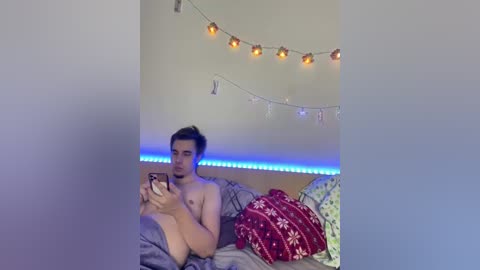 Video of a shirtless young man with fair skin, medium build, and dark hair, lying on a bed with gray sheets. He holds a phone, surrounded by festive decorations and string lights.