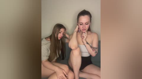 Video of two young women with fair skin, one with long brown hair, one with a ponytail, sitting on a gray couch, touching each other's feet, wearing casual attire.