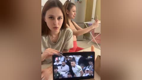 Video of two young women watching TV in a kitchen; one wears a gray top, the other a red tank top. The TV screen shows a news broadcast.