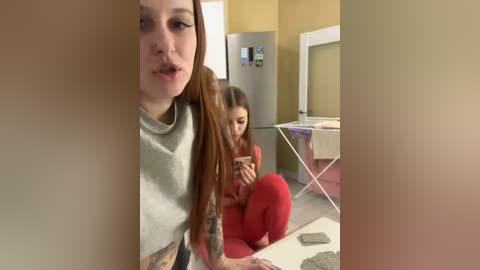 A video of a young woman with long dreadlocks and tattoos, wearing a gray shirt, sitting on a table in a kitchen. Behind her, a young girl in red pajamas is focused on her phone.