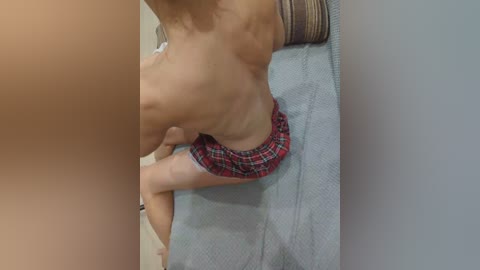 A video of a topless woman with fair skin, wearing a red plaid skirt, sitting on a grey couch in a dimly lit room.