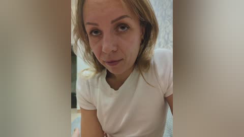 A video of a young woman with light skin and straight, shoulder-length blonde hair, wearing a white T-shirt. She gazes directly at the camera with a neutral expression. The background shows a blurry, beige-colored room.