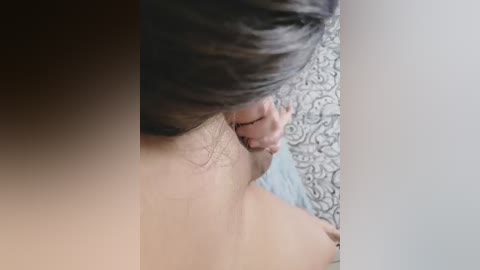 A close-up video shows a person's bare back and shoulder, with dark hair, facing a white wall. The background features a light blue carpet with a subtle floral pattern.