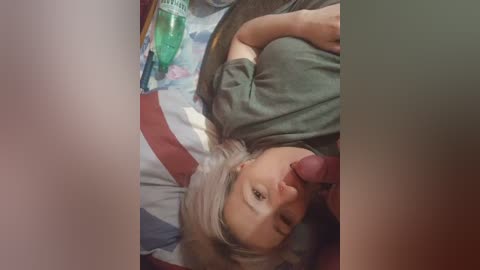 A video shows a young blonde woman lying on a bed, with a man's erect penis in her mouth, surrounded by a disorganized room with a green bottle.