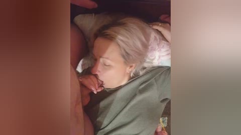 Video of a blonde woman with closed eyes, lying on her side, performing oral sex on a man. She is partially covered by a green blanket. The image is framed by a blurred, reddish-brown border.