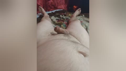 Video of a naked, light-skinned, overweight man lying on a bed, legs spread, with visible pubic hair and flaccid penis. Background includes a red blanket, floral bedspread, and a black pillow.