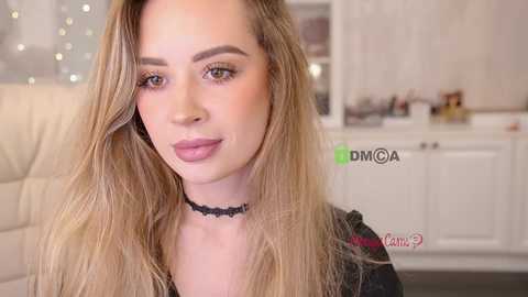 Video of a fair-skinned, blonde-haired woman with long, wavy hair, wearing a black lace choker and black top, smiling softly in a bright, modern kitchen with white cabinets and a cream-colored chair.