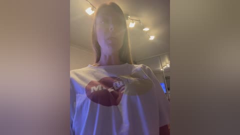 Video of a young woman with long, straight brown hair, wearing a white Hooters T-shirt, standing in a dimly lit room with hanging lights and a blurred background.