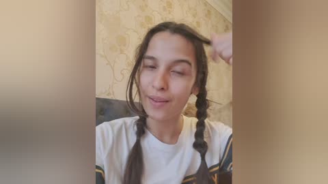 Video of a young woman with long dark hair in two braids, wearing a white shirt with black accents, smiling with eyes closed, adjusting a braid, against a beige, patterned wallpaper background.