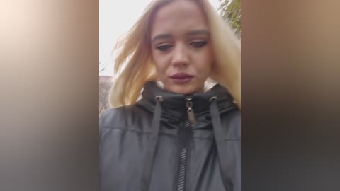 Video of a young woman with long, blonde hair, wearing a black raincoat with a high collar, standing in a blurred outdoor setting with trees in the background.