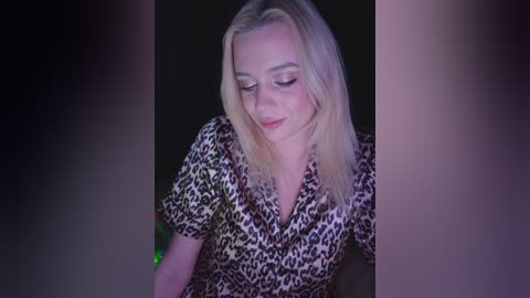 A video of a blonde woman with fair skin, wearing a leopard-print blouse, smiling gently, her eyes closed, against a dark background.