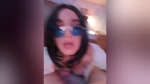 A video shows a close-up of a fair-skinned woman with straight, shoulder-length black hair and blue reflective sunglasses, leaning forward. She's wearing a white top. The blurred background suggests an indoor setting.