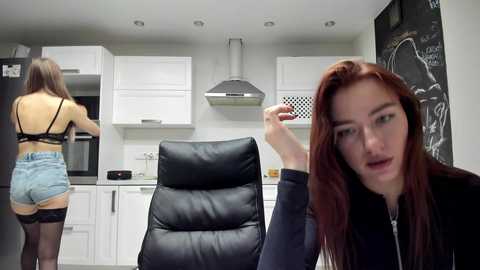 Video of a modern kitchen with a white countertop, black leather chair, and two young women, one with auburn hair and another with brown hair, wearing black tops and denim shorts.