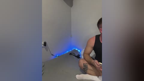 A video of a muscular man with short hair, wearing a black tank top, sitting cross-legged on a gray mat in a bare, white-walled room. He has a tattoo on his left thigh. Blue LED lights are installed along the walls, casting a cool glow.