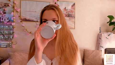 A video of a blonde woman with fair skin drinking from a white cup, surrounded by pink cherry blossom garlands, a Paris-themed poster, and a beige sofa.