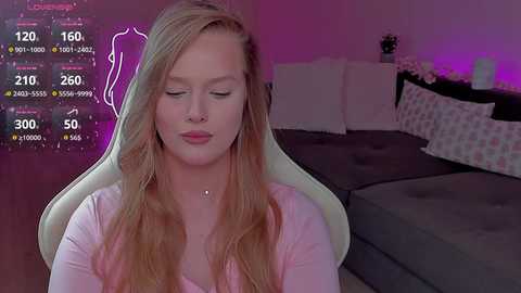 Video of a blonde, fair-skinned woman with long hair, wearing a pink top, sitting on a white gaming chair in a dimly lit bedroom with a bed and floral decorations.