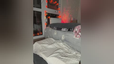 A video of a cozy bedroom with a gray bedspread, white duvet, and plush toys. Red glowing lights adorn the wall, creating a warm ambiance. A radiator and window with curtains are visible in the background.