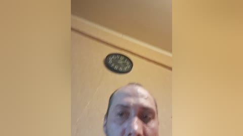 Video of a bald, middle-aged man with a light complexion, standing against a mustard-yellow wall. A circular black and silver ceiling fixture is centered above him. The image is slightly blurred.