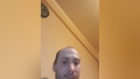 Video of a bald, light-skinned man with a mustache, wearing a dark shirt, standing in a yellow-tiled bathroom with a metallic showerhead and a visible wall-mounted light fixture.