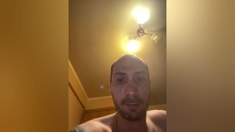 A close-up video of a shirtless, bald man with a beard, standing indoors. The background features a warm-toned ceiling with two bright light bulbs and a wooden ceiling trim.