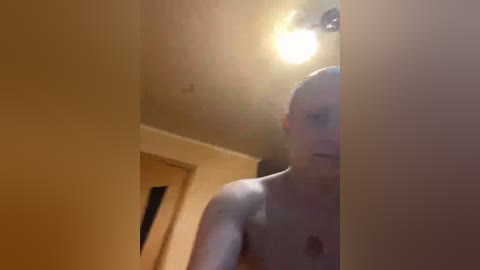 A blurry video of a bald, shirtless man with a light complexion, captured from a low angle, standing in a dimly lit, beige-walled room with a small, lit ceiling light.