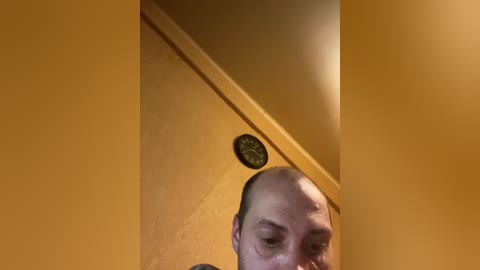 A video of a bald man with a light complexion and visible sweat on his face, standing in a dimly lit room with yellow walls and a round clock on the ceiling.