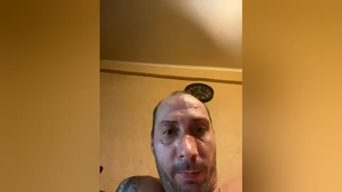 Video of a bald, light-skinned man with a mustache, standing shirtless against a mustard-yellow wall, with a ceiling fan visible overhead.