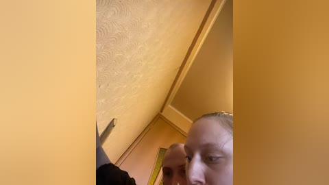 A video of a close-up, low-angle shot of a person with light skin, short hair, and a yellow measuring tape near their mouth. The background features textured beige walls and a wooden door frame.