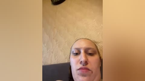 A video of a Caucasian woman with light skin and straight, blonde hair, lying on a grey couch, partially visible. The background shows a beige textured wall with a subtle floral pattern.