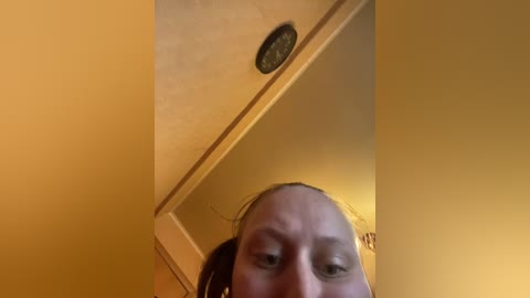 Video of a woman with light skin, dark hair, and brown eyes, taken from a low angle, showing her face and part of her reflection in a mirror. The background is a beige wall with a small circular light fixture.