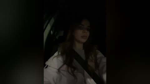 A dimly-lit video shows a woman with long, wavy hair, wearing a light-colored coat, sitting in a car at night. She appears to be in a reflective, contemplative state.