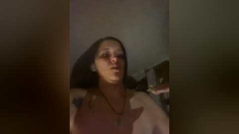 Video of a topless woman with medium-length brown hair, wearing a gold necklace, in a dimly lit room with a blurred background.