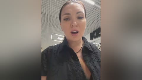 Video of a light-skinned woman with dark hair pulled back, wearing a black lace blouse, mouth open in an expression of surprise or shock, in a tiled, modern room.