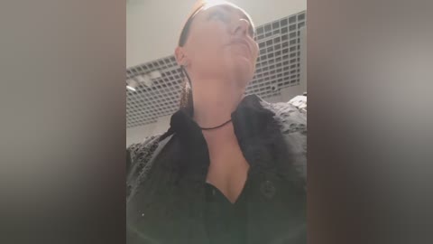 Video of a woman with light skin and brown hair, wearing a black robe, standing in a tiled bathroom with a grid-patterned ceiling. She gazes upward, head tilted back, with a serene expression.