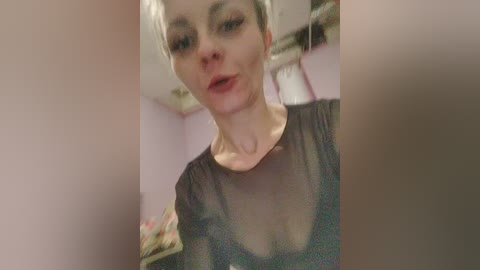 A blurry video of a woman with short blonde hair, fair skin, and a black sheer top, standing indoors with pink walls and ceiling fan in the background.